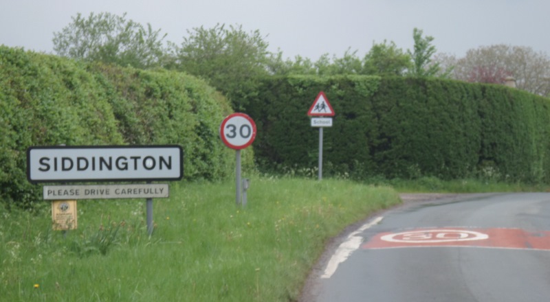 Entrance from the main road to Siddington village.  To the left of the road is a wide green verge and hedgerow. There are three signs on the grassy verge, one to acknowledge the start of Siddington, a 30 mile per hour limit sign and a warning to watch out for school children