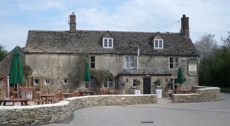 A front-on view of the Greyhound Pub in Siddington, Gloucestershire. Its early spring with blue skies. The pub is made of grey stone and a slate roof. There is an outdoor seating area at the front with wooden benches and tables and dark green parasols. A small dry-stone wall surrounds the garden 